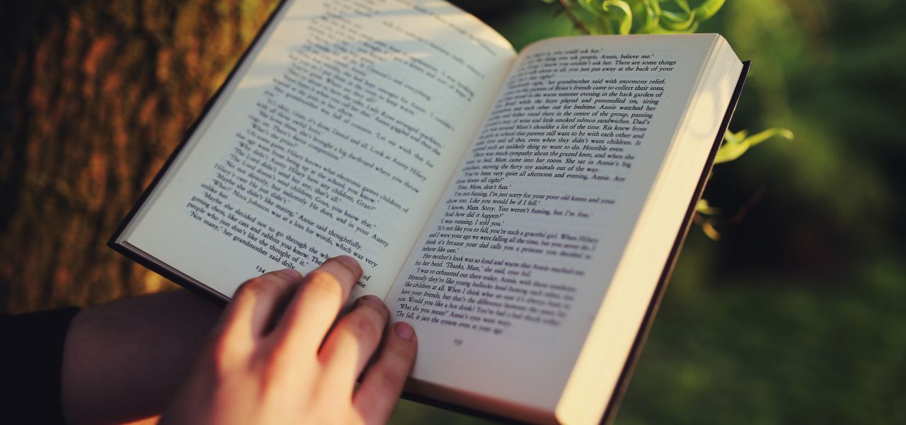 An open book on someone's lap as they read beneath a tree on a sunny day
